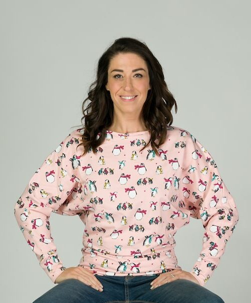 Dark haired lady sitting on a stool with hand on hips wearing a pink top with penguins